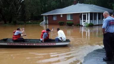 Firefighters use a rowboat to transport a man to safety. In the background, brown flood waters surround a one-story brick home.