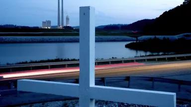 A white cross stands next to a highway with a power plant in the background