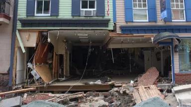 The ground floor of a building is completely gutted and the front wall is missing, with debris lying on the street in front of it.