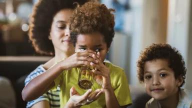A child drinking water out of a glass an and adult behind them holding up a hand below the glass