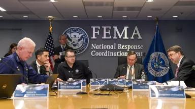 Five men sit at a table under a sign that says “FEMA National Response Coordination Center.”