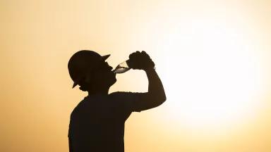 A person with a round hat drinking a bottle of water with the sun in the background