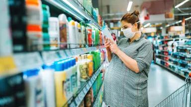 A pregnant woman wearing a face mask reads the ingredients on a bottle in a grocery store aisle