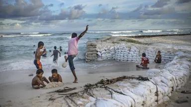 Children play on a beach in front of a long row of stacked sandbags