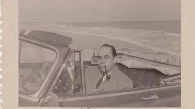 A black-and-white photo of a man sitting in a convertible on the beach, smoking a pipe. The name "Enrique" is written in the margin of the photo.