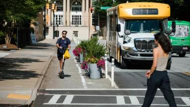 A man rides a scooter in a protected bike lane in Atlanta, while a woman crosses the street and a University shuttle approaches 