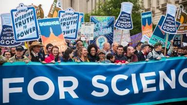 People march at a protest behind a large banner and holding signs that read "Science speaking truth to power" and "Science serving the common good"