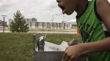 A child drinks from a water fountain in a park, with an apartment building visible in the background