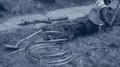 A black and white image of a worker in a trench dug next to a sidewalk with a segment of coiled metal pipe next to him