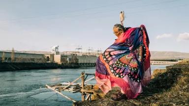 A woman wearing a colorful shawl with the image of a butterfly on it stands on a riverbank