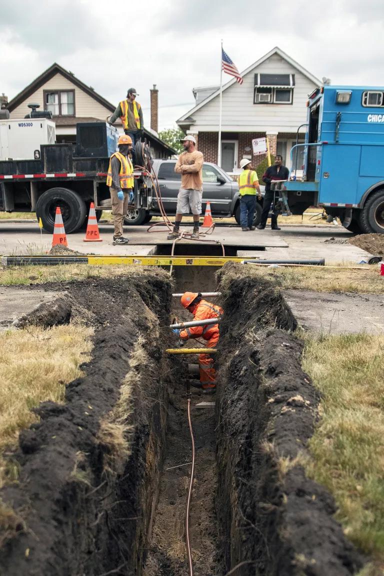 A worker stands in a deep trench dug into the front yard of a house, with more workers and large trucks in the street in the background.