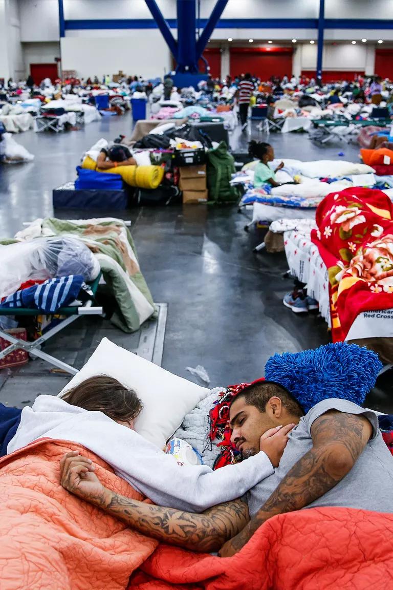 A man and woman rest under blankets on cots on a large convention center floor