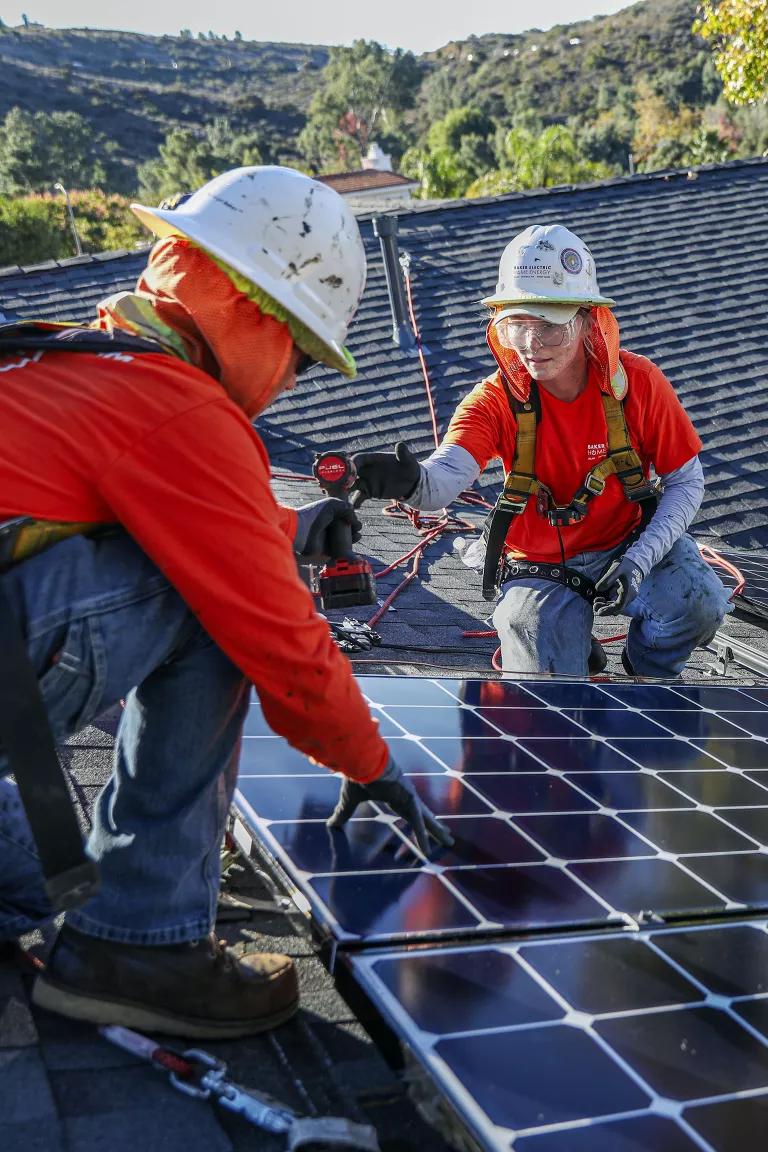 Two solar installers wear safety equipment secure panels to a pitched roof. 