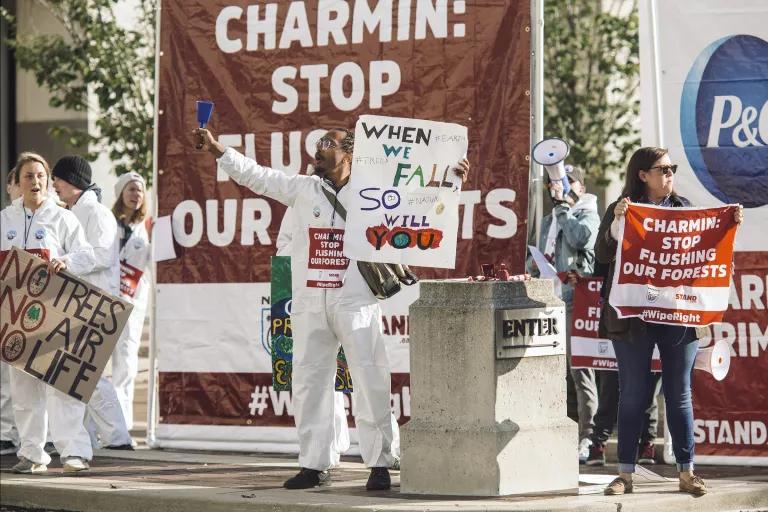 A group of protesters hold signs in front of a large banner reading "Charmin: Stop Flushing Our Forests"