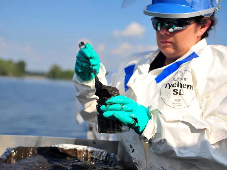 A woman in protective gear pours a thick black sludge into a clear container.